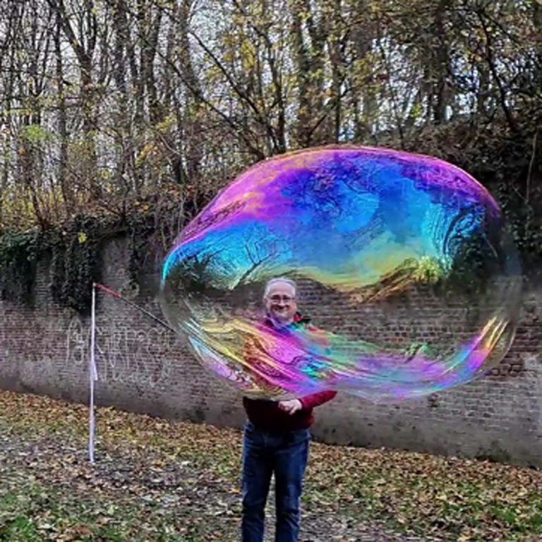 Een man met een grote, kleurrijke zeepbel staat buiten tussen de bomen.