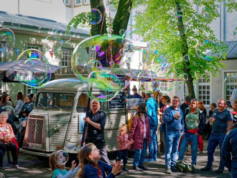 Mensen vieren feest buiten terwijl zeepbellen rond een oude wagen zweven.