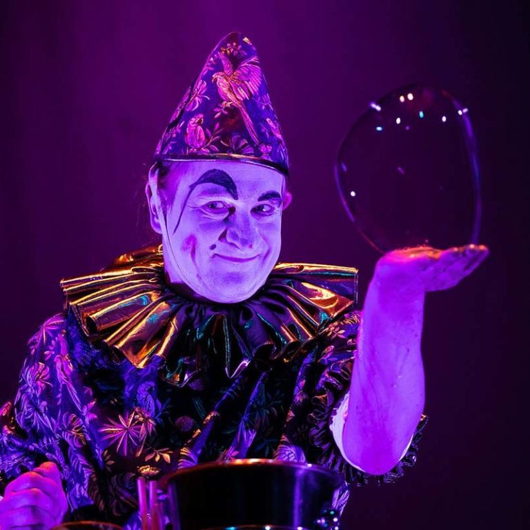Een clown in een kleurrijk kostuum houdt een grote zeepbel in zijn hand.
