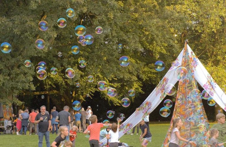Kleurrijke zeepbellen zweven boven een speelplaats met een tent en spelende kinderen.